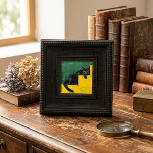 Op de afbeelding: Een ingelijst kunstwerk toont een zwarte kat die rust op gele trappen tegen een groene achtergrond. De vierkante lijst is zwart en heeft een textuurontwerp. Het kunstwerk wordt tentoongesteld op een houten oppervlak, met antieke boeken en een vergrootglas op de achtergrond.