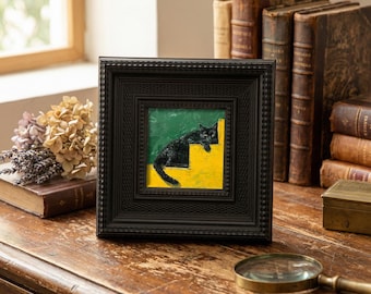 May include: A black framed artwork shows a black cat on yellow steps against a green backdrop. The square frame is black with a textured pattern. The artwork is on a wooden surface, with antique books and a magnifying glass in the background.