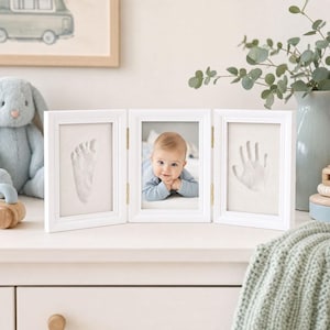 May include: A white, three-panel hinged picture frame featuring a central photograph of a baby in a blue shirt, flanked by two side panels containing white, raised impressions of a single baby footprint and a single baby handprint.