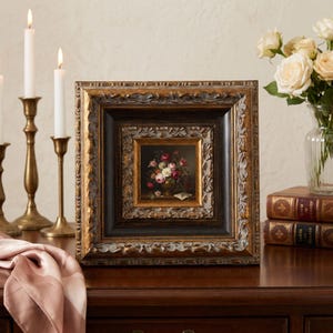Puede incluir: Un cuadro enmarcado con un bodegón floral, con un jarrón de rosas rosas, rojas y blancas. El marco ornamentado es dorado y negro. El cuadro está sobre una superficie de madera oscura, con velas y libros en el fondo.