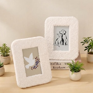 Könnte beinhalten: Zwei cremefarbene, strukturierte Fotorahmen. Einer zeigt eine Taube mit einem Beerenstrauß, der andere eine Katzen- und Hundeillustration. Die Rahmen sind rechteckig mit abgerundeten Kanten und stehen auf einer Holzoberfläche.