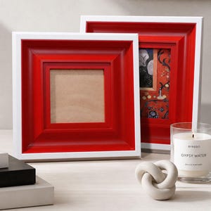 May include: Two square picture frames with a red inner frame and a white outer frame. One frame is empty, and the other contains a colorful image. A candle and decorative knot are also visible.