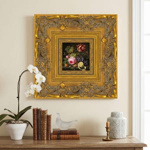 May include: A framed still life painting featuring a vibrant arrangement of grapes, a pink rose, and a tulip. The painting is set within a gold ornate frame, mounted on a cream-colored wall. A white orchid, books, and decorative items are on a wooden surface below.