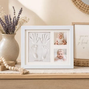 May include: A white framed baby keepsake with handprints, footprints, and two baby photos. The frame includes the name "Penelope" and is displayed on a wooden surface with a vase of lavender.