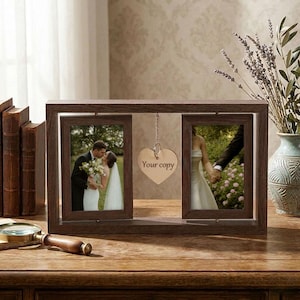 May include: A dark brown wooden double photo frame with two wedding photos. A heart-shaped charm with the text "Your copy" hangs between the frames. The frame sits on a wooden surface.