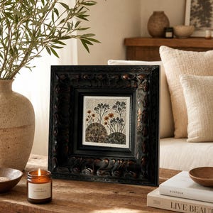 Könnte beinhalten: Ein gerahmtes Kunstwerk mit einem schwarzen, verzierten Rahmen. Das Kunstwerk zeigt ein botanisches Design mit stilisierten Bäumen und Blumen in Orange-, Braun- und Grüntönen. Eine brennende Kerze in einem Glas und Bücher stehen auf dem Holztisch.