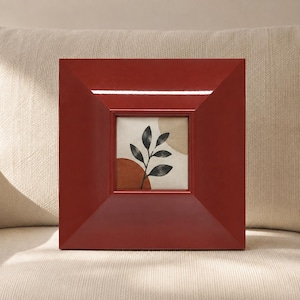 May include: A square, deep red picture frame with a wide, angled border. The frame holds a print featuring a black botanical leaf illustration centered over abstract shapes in muted tan and burnt orange colors on a cream-colored background.