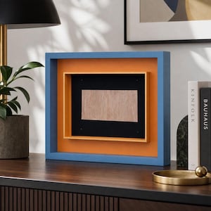 May include: A square, layered picture frame with a blue outer frame, orange middle frame, and black inner frame. The center features a light wood-grain rectangle. The frame sits on a dark wood surface.