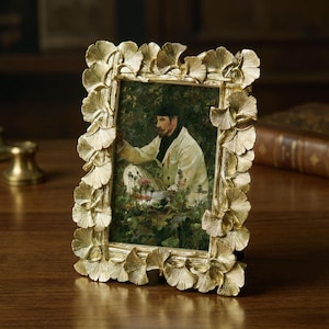 以下が含まれることがあります： イチョウの葉のデザインが施された金色のフォトフレーム。フレームには、白いコートと黒いスカーフを着た男性の絵画が収められています。フレームは木製の表面にあります。