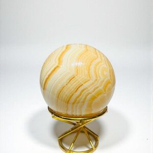 May include: A decorative sphere with a layered, cream and amber pattern, resting on a gold-colored metal stand. The sphere's design resembles natural stone or agate. The stand has a geometric, openwork design.