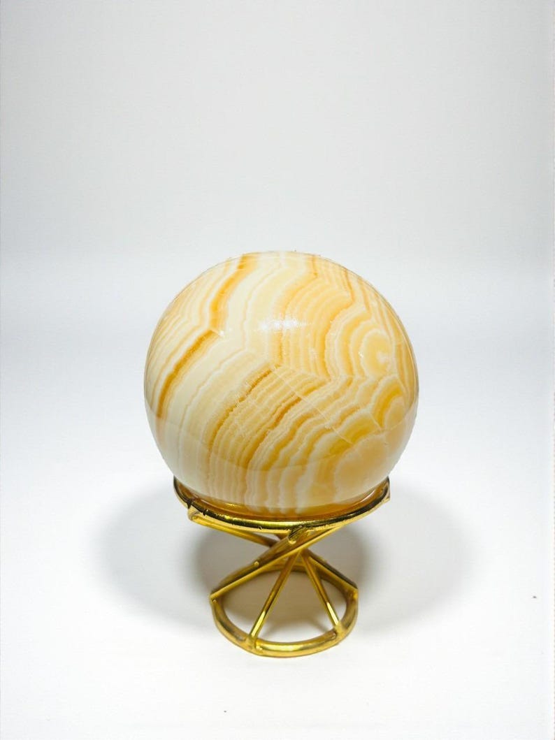 May include: A decorative sphere with a layered, cream and amber pattern, resting on a gold-colored metal stand. The sphere's design resembles natural stone or agate. The stand has a geometric, openwork design.