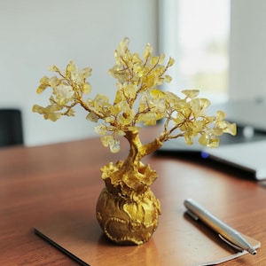 Puede incluir: Una escultura decorativa de árbol con una base dorada y ramas adornadas con piedras amarillas en forma de cristal. El árbol está sobre una superficie de madera, junto a un bolígrafo y una placa de color cobre. Es un artículo de decoración único.