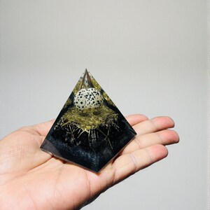 Op de afbeelding: Een handgemaakt piramidevormig kunstwerk van hars. De piramide heeft een gevlekt eivormig object genesteld in een gouden nestachtige structuur, ingekapseld in heldere hars met een zwarte basis. Het kunstwerk wordt in een hand gehouden.