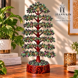 May include: A decorative tree sculpture featuring a glossy dark red trunk and base, adorned with numerous small, polished green and purple crystal beads attached to thin gold-toned wire branches. The text HASNAIN CRYSTALS Elevate Your Space is visible.
