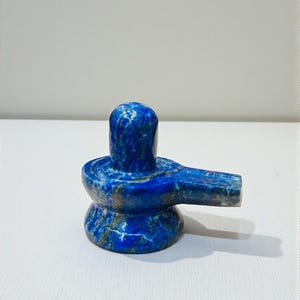 May include: A blue stone lingam sculpture, a symbol of Lord Shiva. The sculpture is crafted from a deep blue stone, featuring lighter blue and grey veining. The lingam has a rounded top and a flat base with a spout.