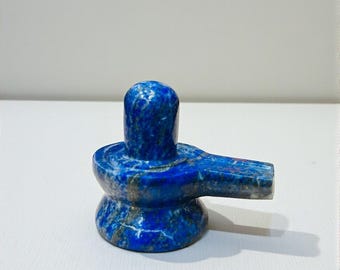 May include: A blue stone sculpture of a lingam, a symbol of Lord Shiva. The sculpture is made of a deep blue stone with lighter blue and gray veining. The lingam has a rounded top and a flat base with a spout.