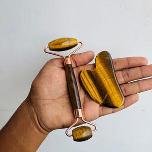 Puede incluir: Un rodillo facial y una herramienta gua sha en forma de corazón de piedra ojo de tigre. El rodillo tiene un mango y un marco de color cobre. La piedra es de color marrón dorado con rayas marrones oscuras. Las herramientas se sostienen en una mano sobre un fondo blanco.