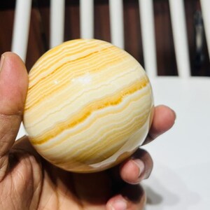 May include: A polished, spherical decorative stone with a banded pattern of creamy white and golden yellow hues. The orb is held in a hand, showcasing its smooth, rounded form. This natural stone sphere is a decorative item.