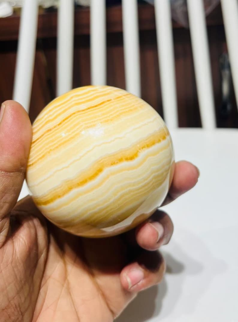 May include: A polished, spherical decorative stone with a banded pattern of creamy white and golden yellow hues. The orb is held in a hand, showcasing its smooth, rounded form. This natural stone sphere is a decorative item.