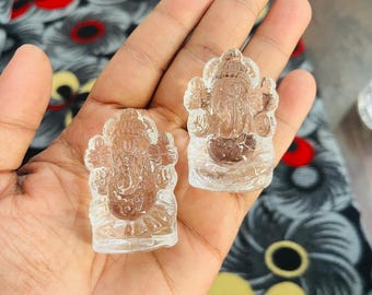 Może przedstawiać: Dwie małe, przezroczyste kryształowe rzeźby hinduskiego bóstwa Ganesha. Rzeźby są misternie wyrzeźbione ze szczegółami boga z głową słonia. Figurki trzymane są w dłoni, z wzorzystą tkaniną w tle.