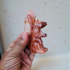 May include: A carved squirrel figurine in shades of red, brown, and white. The squirrel is in a sitting position with its tail upright. The figurine is made of a stone-like material.
