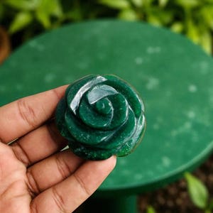 Könnte beinhalten: Eine geschnitzte, dunkelgrüne Steinrose. Der Edelstein wird in einer Hand gehalten, mit einem verschwommenen grünen Hintergrund aus Blättern und einer grünen Oberfläche. Die Rose hat detaillierte Blütenblätter und eine polierte Oberfläche.