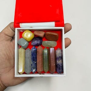 Könnte beinhalten: Eine weiße Box mit rotem Innenraum, die eine Sammlung polierter Edelsteine und Kristallstäbe enthält. Die Edelsteine sind in verschiedenen Farben, darunter Gelb, Lila und Grün. Die Kristallstäbe sind vertikal und mehrfarbig.