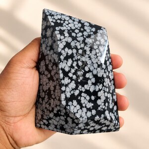 Pode incluir: Uma pedra de obsidiana floco de neve polida com uma base preta e padrões radiais brancos em forma de flor. A pedra é cortada numa forma geométrica multifacetada com arestas afiadas e uma superfície lisa e refletora em toda a peça.
