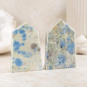 Puede incluir: Dos tallas de piedra en forma de casa con acabado pulido. Las piedras muestran un patrón moteado de color crema claro, verde pálido e inclusiones minerales de color azul vibrante con pequeñas motas gris oscuro en la superficie.