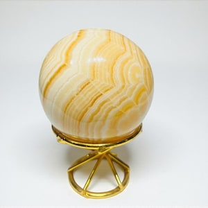 May include: A decorative sphere with a swirling pattern of cream and amber hues, displayed on a gold-colored metal stand. The sphere's polished surface reflects light, and the stand has a geometric, open design.