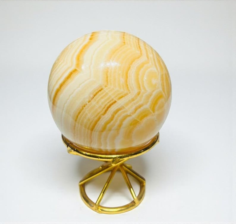 May include: A decorative sphere with a swirling pattern of cream and amber hues, displayed on a gold-colored metal stand. The sphere's polished surface reflects light, and the stand has a geometric, open design.