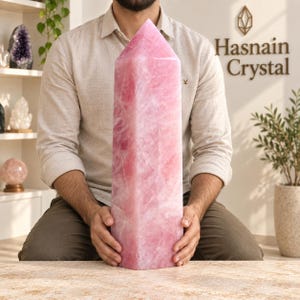 May include: A large, polished rose quartz crystal tower with a hexagonal base and a pointed top. The stone features a translucent, pale pink colour with natural internal fractures and cloudy white inclusions throughout its solid, vertical structure.