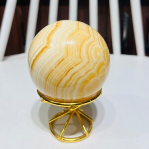 May include: A polished, spherical decorative object with a swirling pattern of cream and amber hues. The sphere rests on a small, gold-colored metal stand with a geometric design. The overall aesthetic is elegant and decorative.