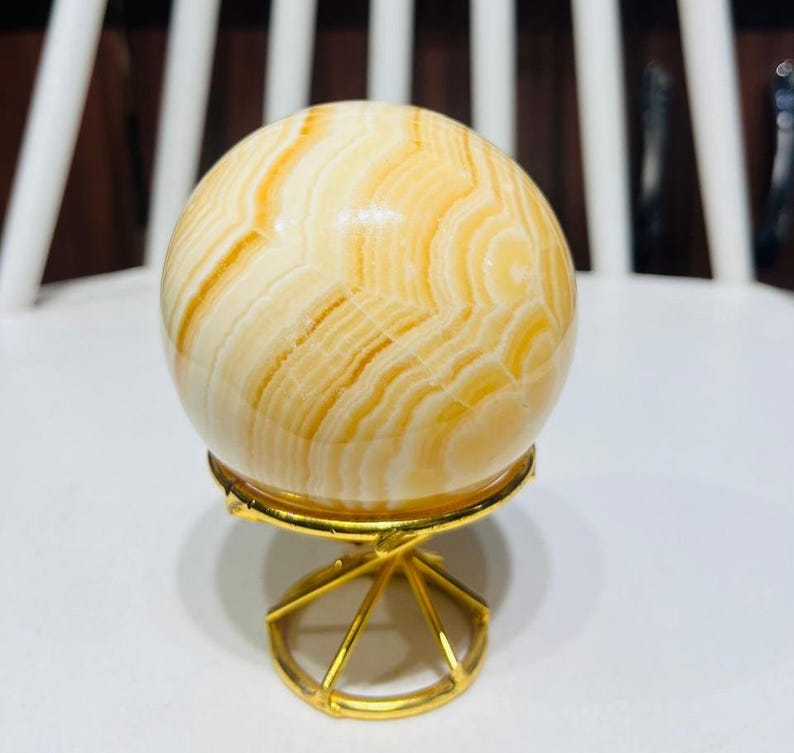 May include: A polished, spherical decorative object with a swirling pattern of cream and amber hues. The sphere rests on a small, gold-colored metal stand with a geometric design. The overall aesthetic is elegant and decorative.