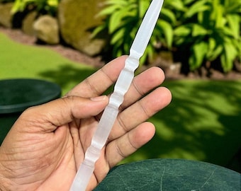 Könnte beinhalten: Ein klarer, länglicher Kristallstab mit einer Spitze und dekorativen, geriffelten Abschnitten. Der Stab wird vor einem Hintergrund aus grünem Laub und einer dunkelgrünen Oberfläche gehalten.