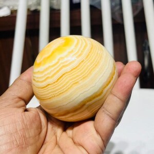 May include: A spherical decorative object with a banded pattern of yellow and cream colors. The orb is held in a hand, showcasing its smooth, polished surface. The object appears to be made of stone or a similar material.