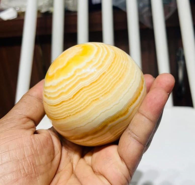 May include: A spherical decorative object with a banded pattern of yellow and cream colors. The orb is held in a hand, showcasing its smooth, polished surface. The object appears to be made of stone or a similar material.