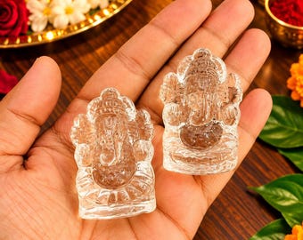 Puede incluir: Dos pequeñas figuras de cristal transparente talladas que representan a la deidad Ganesha. Cada figura presenta detalles intrincados de la figura con cabeza de elefante sentada en postura meditativa, con una superficie lisa, translúcida y reflectante.