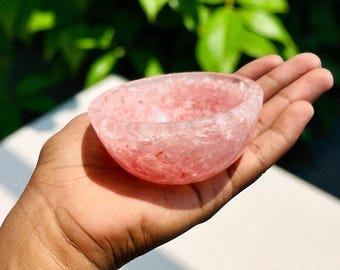 May include: A small, pink crystal bowl with a rough, textured surface. The bowl is held in a hand, with a blurred green leafy background. The bowl is likely used for decorative purposes or for holding small items.