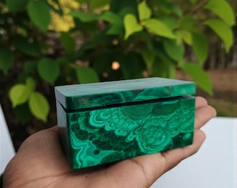 May include: A small, rectangular box made of polished malachite, a vibrant green stone with swirling patterns. The box has a hinged lid and is held in a hand, showcasing the stone's natural beauty. The box is a decorative item.