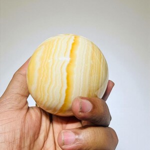 May include: A polished, spherical decorative object with a warm, yellow and cream color palette. The sphere has a banded pattern with vertical lines of varying shades of yellow and cream. The object is held in a hand.
