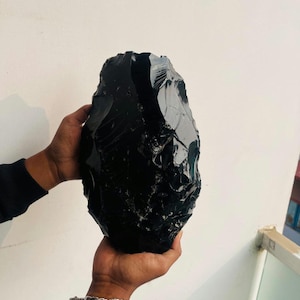 Peut inclure: Une grande pierre d'obsidienne de forme irrégulière, tenue à deux mains. La pierre est d'un noir profond et brillant avec une surface rugueuse et texturée. Les mains tiennent la pierre devant un mur blanc uni.