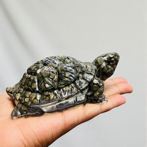 Puede incluir: Una detallada figura de tortuga de piedra tallada en tonos grises y dorados. La tortuga se sostiene en una mano, mostrando los intrincados detalles del caparazón y el cuerpo. La figura tiene una apariencia texturizada, lo que sugiere un material de piedra natural.