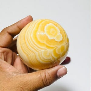 May include: A polished, spherical decorative object with concentric bands of yellow and white. The orb is held in a hand, showcasing its smooth, patterned surface. The overall theme is a decorative item.