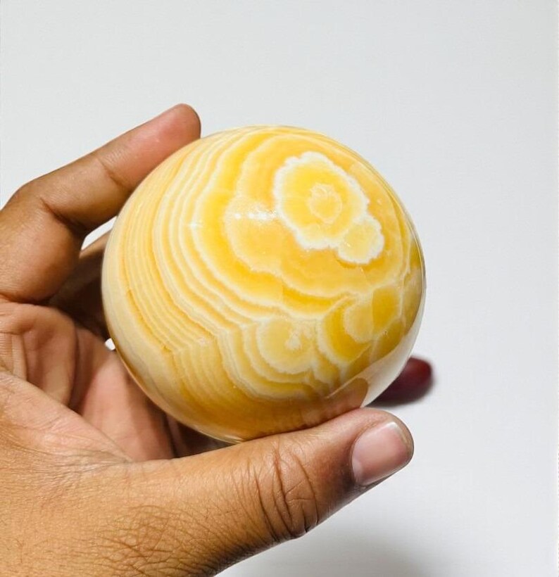 May include: A polished, spherical decorative object with concentric bands of yellow and white. The orb is held in a hand, showcasing its smooth, patterned surface. The overall theme is a decorative item.