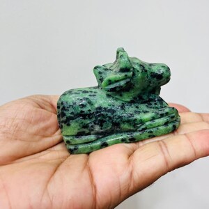 Puede incluir: Una pequeña figura de piedra tallada a mano que representa una vaca o un toro en reposo. La escultura está hecha de un mineral moteado en verde y negro, con una superficie lisa y pulida que resalta las variaciones de color naturales.