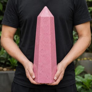 Peut inclure: Une grande tour en cristal à six côtés avec un sommet pointu, présentant une couleur rose poussiéreux unie et une surface mate et texturée. L'objet a la forme d'un obélisque avec des faces verticales plates qui se rejoignent en un sommet aigu.