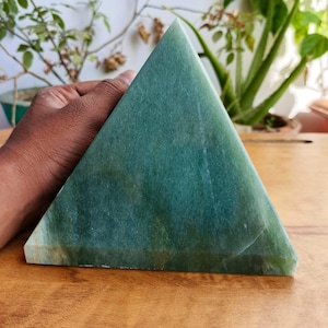 Puede incluir: Una piedra verde sólida en forma de pirámide con una superficie lisa. La pirámide está sostenida por una mano y descansa sobre una superficie de madera. La piedra tiene variaciones de color, con tonos verdes más claros y oscuros.