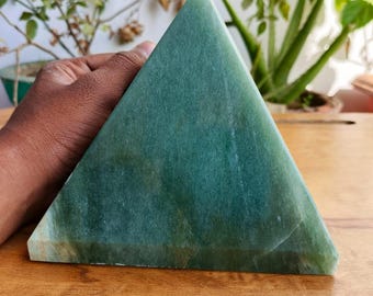May include: A solid, green pyramid-shaped stone with a smooth surface. The pyramid is held by a hand and sits on a wooden surface. The stone has variations in color, with lighter and darker green hues.