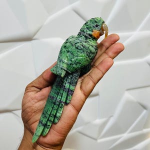 以下が含まれることがあります： 複雑な羽のディテール、長い尾、滑らかな明るい色のくちばしが特徴の、緑色の石で彫られたオウムの置物。彫刻全体に自然な暗い斑点と深い緑色の色合いが見られ、鳥が休んでいる姿勢を捉えています。